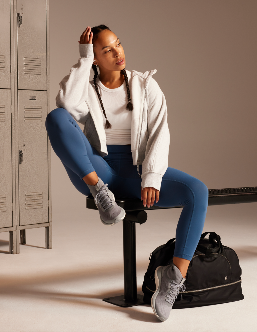 lululemon locker room — model in teal leggings, grey hoodie, bench and metal lockers, gym bag